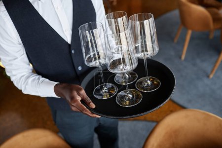Téléchargez les photos : Gros plan d'un serveur classique tenant un plateau avec des verres à vin en cristal tout en mettant des tables dans un restaurant de luxe, espace de copie - en image libre de droit