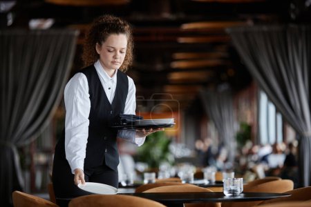 Téléchargez les photos : Portrait taille haute de la jeune femme comme table de réglage de serveur féminin dans un restaurant de luxe se préparant pour l'ouverture, espace de copie - en image libre de droit