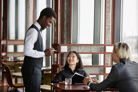 Retrato de vista lateral del joven negro como servidor que recibe órdenes de la pareja y sonríe en un restaurante de lujo, espacio para copiar