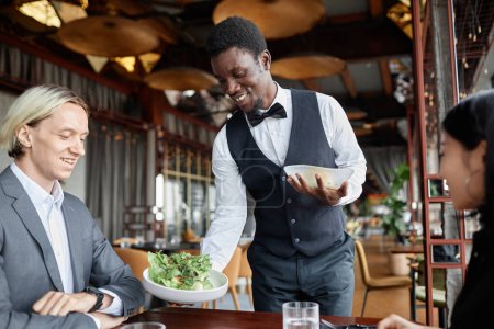 Téléchargez les photos : Portrait de jeune homme noir comme serveur apportant de la nourriture sur plateau pour un couple profitant d'un dîner dans un restaurant de luxe - en image libre de droit