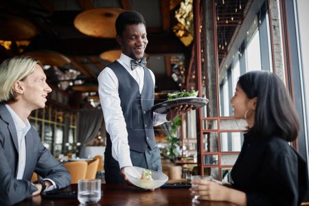 Téléchargez les photos : Portrait de serveur souriant apportant de la nourriture sur le plateau aux invités profitant du dîner dans un restaurant de luxe - en image libre de droit