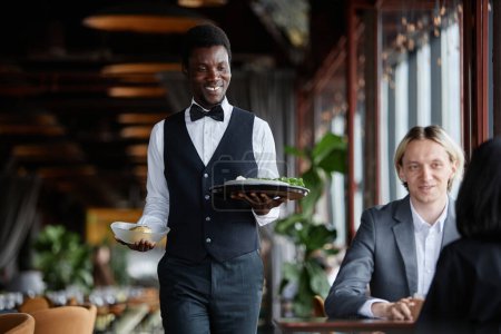 Téléchargez les photos : Portrait de jeune homme noir comme serveur apportant de la nourriture sur le plateau aux invités profitant du dîner dans un restaurant de luxe - en image libre de droit