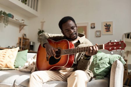Retrato de vista frontal de un joven negro tocando la guitarra acústica mientras disfruta del tiempo libre en una acogedora casa, espacio para copiar