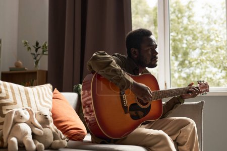 Foto de Retrato de vista lateral del hombre negro adulto tocando la guitarra en casa acogedora con tonos tierra calma, espacio de copia - Imagen libre de derechos