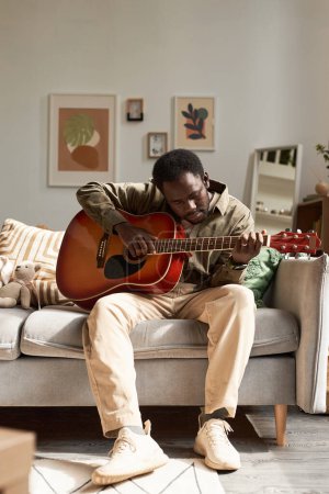 Retrato vertical de cuerpo entero del hombre negro adulto tocando la guitarra en casa iluminado por la luz del sol