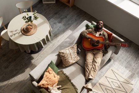 Foto de Vista de arriba hacia abajo del joven negro tocando la guitarra acústica tumbado en el sofá en el acogedor hogar iluminado por la luz del sol, espacio de copia - Imagen libre de derechos