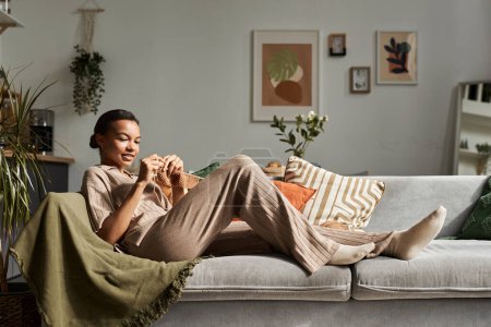 Elegante retrato de larga duración de la joven mujer negra tejiendo en casa mientras se relaja en el sofá en el interior acogedor, espacio de copia