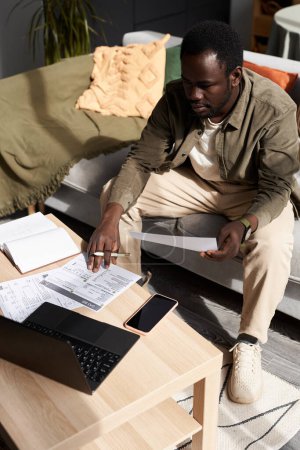 Photo for High angle portrait of adult Black man using laptop while doing taxes and home and calculating budget - Royalty Free Image