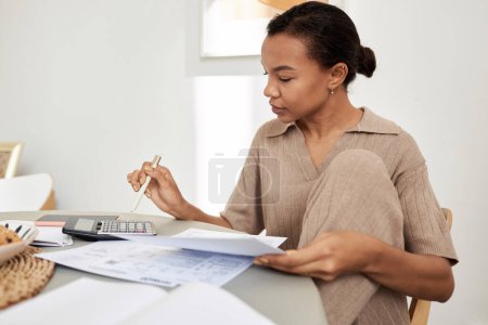 Retrato de vista lateral mínima de la joven mujer negra haciendo impuestos en casa y usando calculadora, espacio para copiar