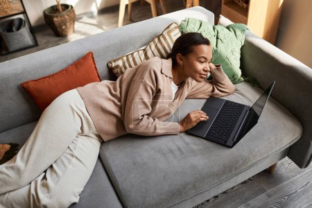 Retrato de ángulo alto de la joven afroamericana acostada en un cómodo sofá en el acogedor hogar iand utilizando el ordenador portátil, espacio para copiar