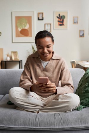 Retrato de larga duración de una chica negra sonriente usando un teléfono inteligente con auriculares inalámbricos mientras navega por las redes sociales sentado con las piernas cruzadas en un acogedor sofá