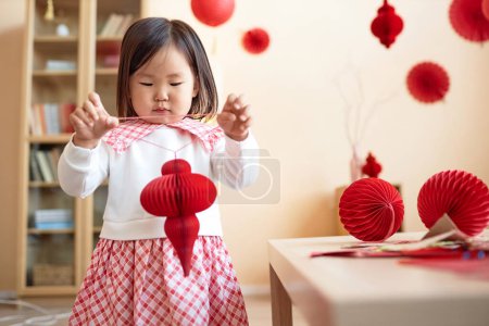 Foto de Cute Asian little girl playing with red paper lanterns and decorations for Chinese New Year celebration at home, copy space - Imagen libre de derechos