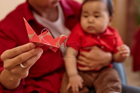 Foto de Primer plano de cuidar padre asiático con lindo bebé sosteniendo grúa de papel rojo a la cámara como símbolo de la celebración de Año Nuevo chino, espacio de copia - Imagen libre de derechos