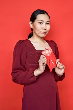 Téléchargez les photos : Portrait vertical de jeune femme tenant une enveloppe hongbao sur fond rouge comme tradition du Nouvel An chinois et regardant la caméra Avoir une abondance débordante chaque année - en image libre de droit