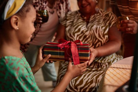 Téléchargez les photos : Grand-mère donnant Kwanzaa cadeau à la petite fille lors de la célébration à la maison - en image libre de droit