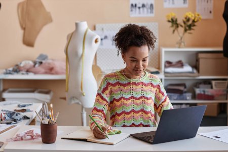 Joven artesana afroamericana viendo videos educativos en línea y tomando notas en copybook mientras está sentada en el lugar de trabajo en una tienda de artesanía