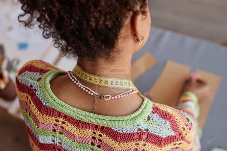 Rückenansicht einer jungen afroamerikanischen Kunsthandwerkerin im handgestrickten Pullover, die am Arbeitsplatz mit Schnittmustern arbeitet