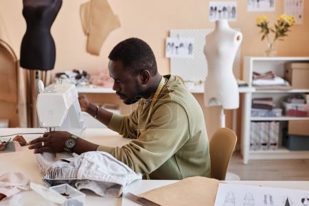 Téléchargez les photos : Vue latérale du jeune artisan afro-américain assis à côté d'une machine à coudre électrique en studio de mode lente et créant une nouvelle tenue - en image libre de droit
