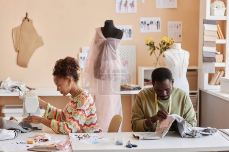 Téléchargez les photos : Deux jeunes créateurs afro-américains ou stylistes cousant de nouveaux vêtements tout en personnalisant des vêtements à l'ancienne en atelier - en image libre de droit