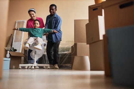 Cheerful young intercultural family of three having fun in living room of new apartment among packed boxes during relocation