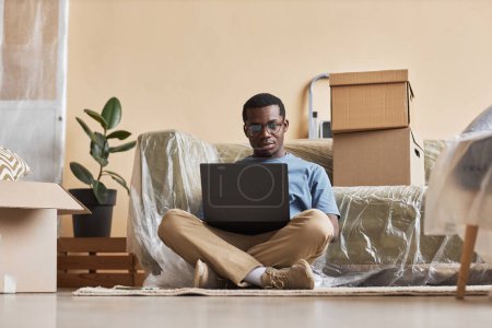Junger seriöser Geschäftsmann mit Brille sitzt auf dem Boden und schaut auf den Laptop-Bildschirm, während er zu Hause vernetzt oder Online-Videos anschaut