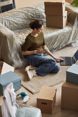 Junge brünette Freiberuflerin mit Laptop, während sie nach dem Umzug in eine neue Wohnung mit gepackten Kartons auf dem Boden des Wohnzimmers sitzt