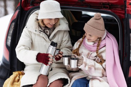 Happy young woman in winterwear pouring hot tea in metallic mugs held by her cute daughter sitting next to her in hatchback of car
