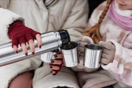 Hands of young woman in mittens pouring hot tea into metallic mugs while sitting by her daughter wearing woolen gloves and winterwear