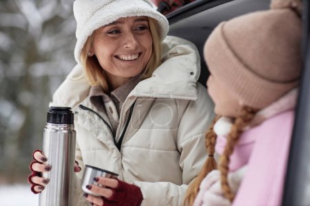 Young smiling mother in winter cap and jacket holding thermos with hot tea and looking at her daughter while both enjoying winter travel