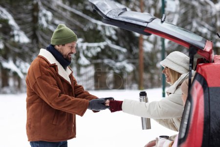 Young cheerful woman in winterwear passing cup with hot tea to her husband standing in front of her during family trip by car