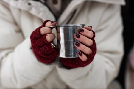 Photo for Focus on hands of young stylish woman in white winter coat and crimson mittens holding metallic mug with hot tea - Royalty Free Image
