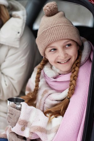 Smiling pretty girl in winterwear looking aside while sitting in hatchback of car next to her mother, enjoying vacation and having hot tea