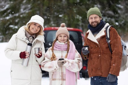 Young happy family of father, mother and daughter with hot tea standing in winter forest with evergreen trees covered with snow