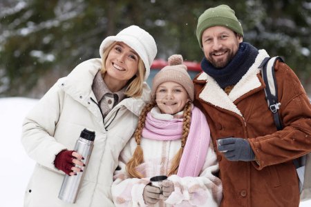 Young cheerful couple and their pretty pre-teen daughter in winterwear looking at camera while standing in winter forest and having tea