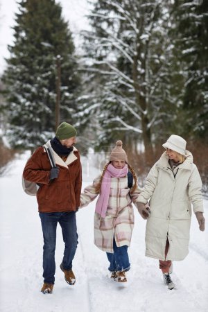 Long shot of young family in winterwear walking down forest road covered with snow while enjoying stroll on winter weekend together