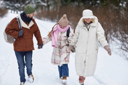 Happy pre-teen girl in winterwear holding by hands of her father and mother while strolling with them along forest road covered with snow