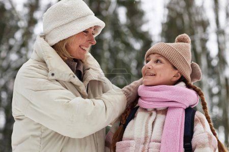 Young cheerful woman in winterwear looking at her adorable pre-teen daughter in warm cap, scarf and coat while chatting to her outdoors