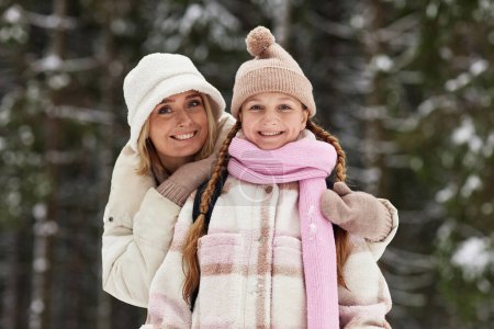 Happy young affectionate mother embracing daughter in winterwear while both standing in winter forest and looking at camera