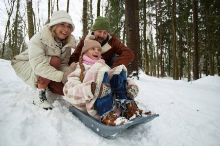 Happy young ecstatic parents pushing their excited pre-teen daughter on sledge down snow hill while playing and having fun in park