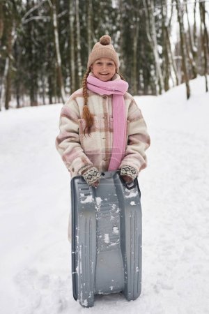 Cute smiling pre-teen girl in winterwear holding toboggan sledge while standing in snowdrift in front of camera and looking at you