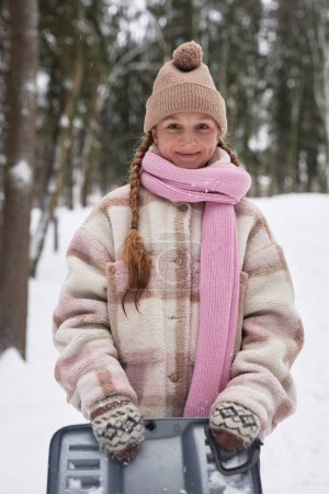 Happy adorable girl in winter coat, cap, scarf and gloves holding toboggan sledge while standing in winter park or forest covered with snow