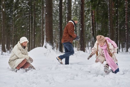 Young mother and daughter rolling snowballs in winter forest or park while man in winterwear moving in snowdrift behind them