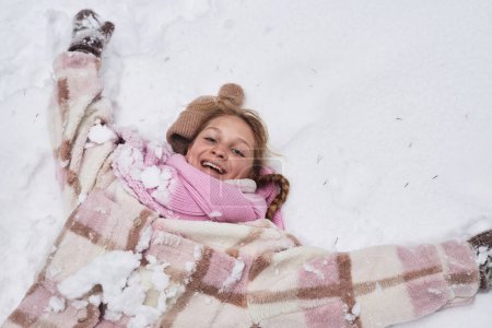 Above angle of youthful cheerful girl in winterwear lying in snowdrift and looking at camera while having fun and enjoying winter
