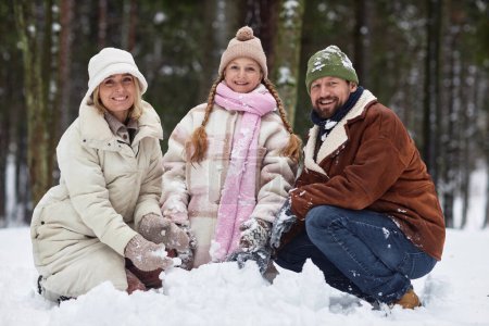 Young cheerful family in winterwear sitting in snowdrift and looking at camera while rolling snowball together and making snowman