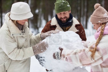 Happy young family of three in winterwear rolling big snowball while making snowman on winter weekend in park or forest