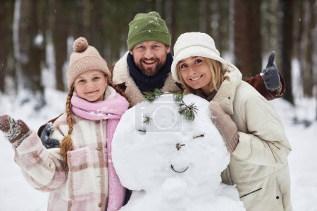 Photo for Cheerful family of three in winterwear standing by snowman in front of camera and looking at you while bearded man showing thumb up - Royalty Free Image