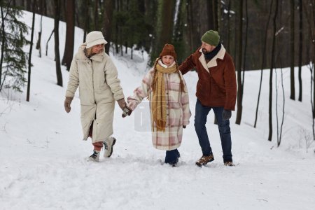 Happy family of three in winterwear walking along forest road covered with snow and chatting while enjoying winter weekend