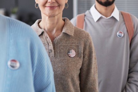 Photo for Mature smiling female voter with USA flag badge on chest standing among other people taking part in presidential elections - Royalty Free Image