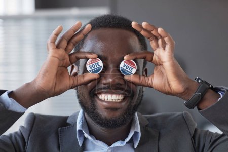 Photo for Happy young African American voter with wide toothy smile holding two small round vote badges by his eyes while standing in front of camera - Royalty Free Image