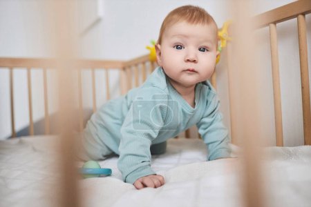 Close up of cute baby boy with blue eyes looking at camera while crawling in crib, copy space
